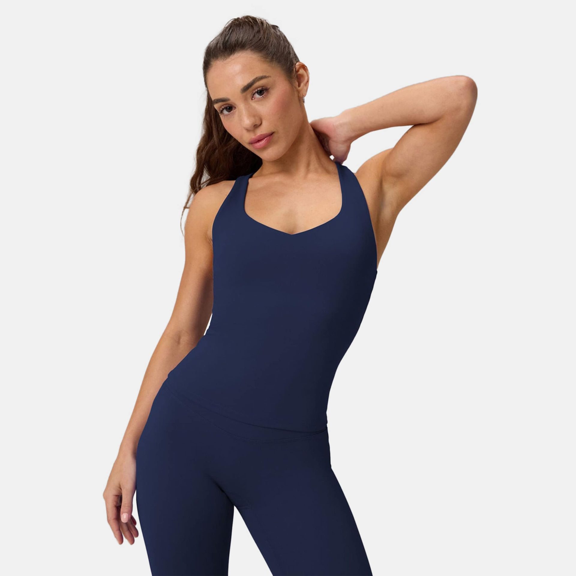 A woman with her hair in a ponytail poses in a dark blue athletic tank top and matching leggings, with one hand resting on the back of her neck.