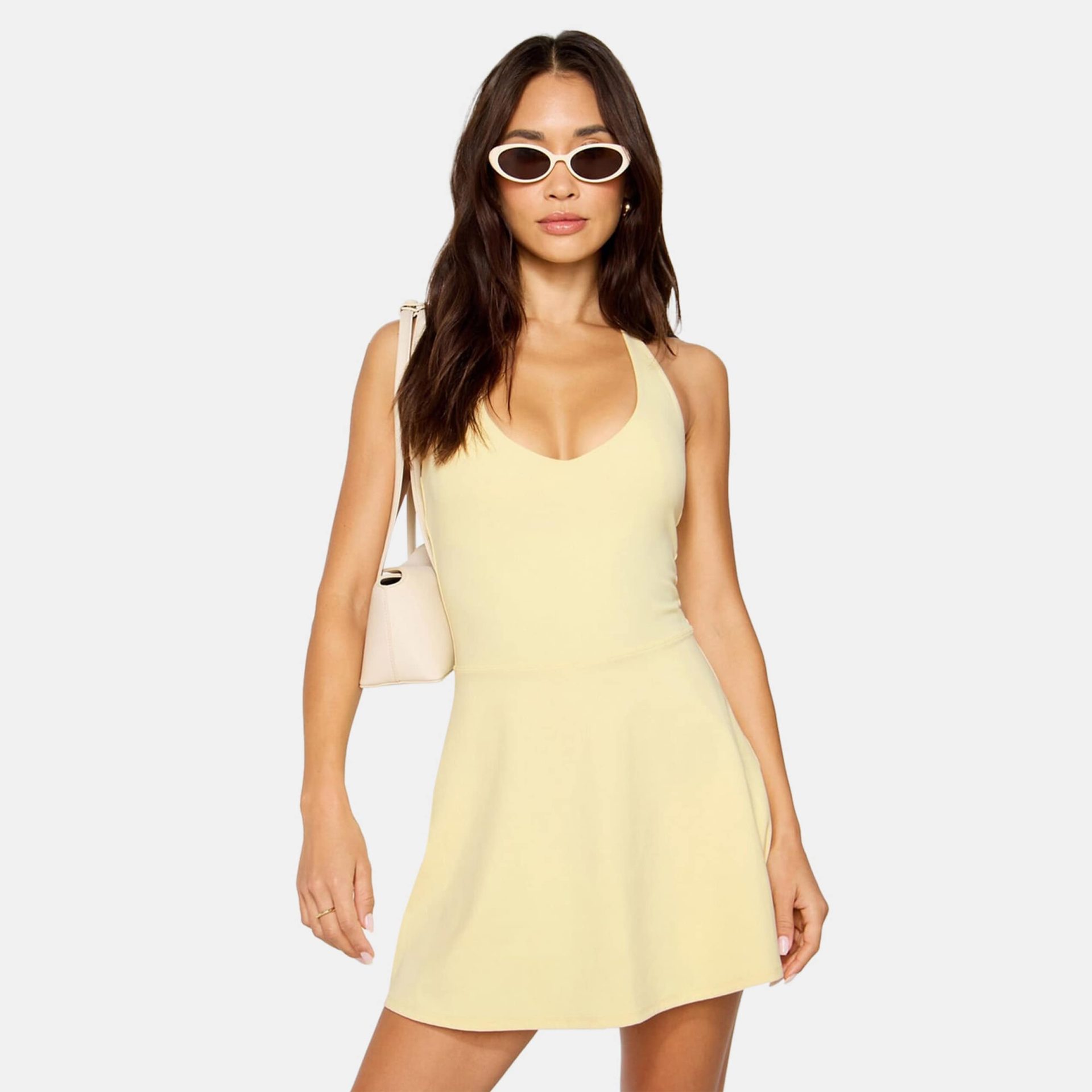 A woman wearing a light yellow halter-style athletic mini dress, white sunglasses, and a small cream shoulder bag.