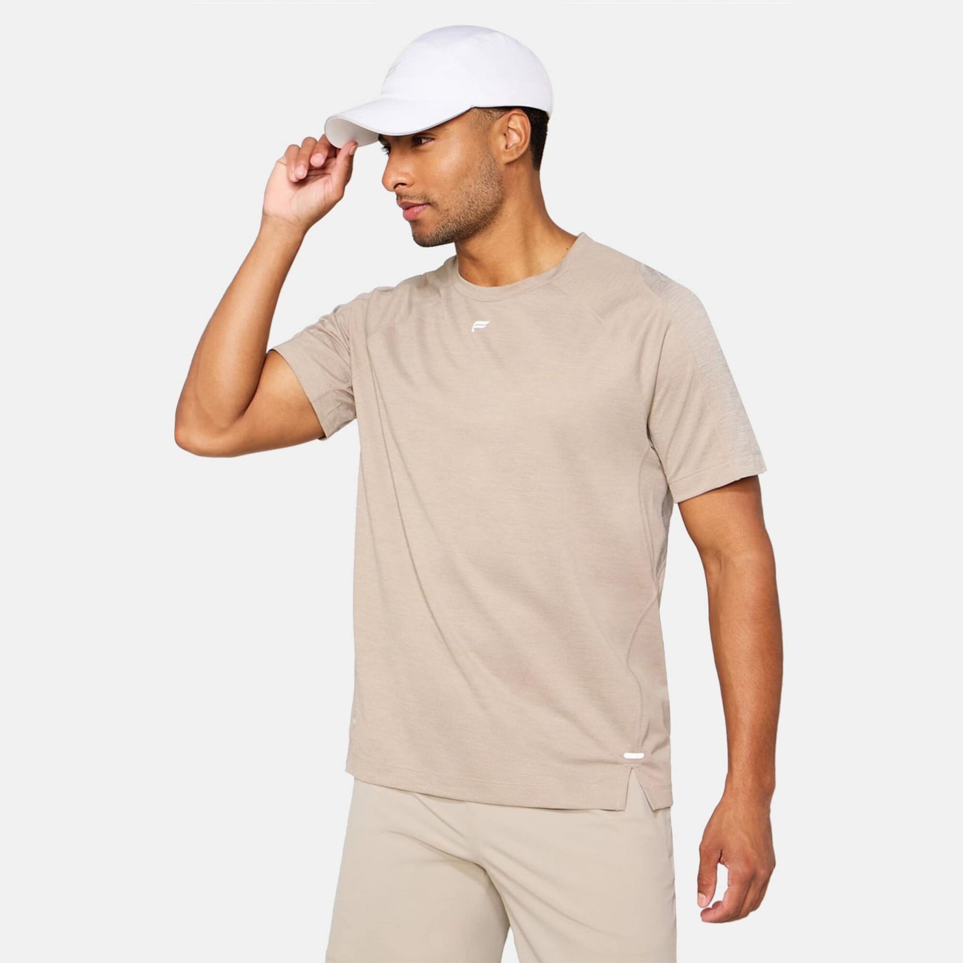 A man wearing a neutral beige t-shirt, matching shorts, and a white baseball cap, adjusting the brim of his hat.